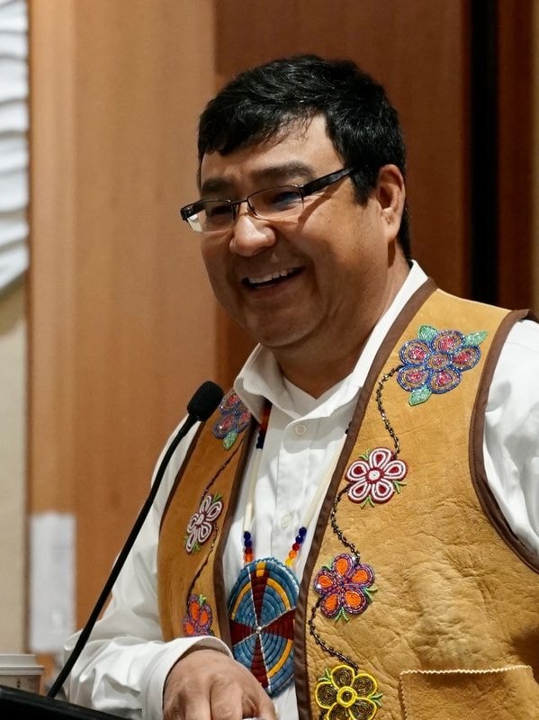 Man smiling while speaking at a microphone, wearing a traditional beaded vest